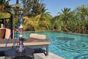 Zona de descanso junto a la piscina con un narguilé sobre una mesa, una toalla enrollada y una piscina cristalina rodeada de vegetación tropical.