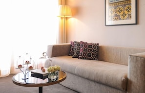 A living room with a beige sofa, patterned pillows, a round coffee table with desserts and flowers, a lamp, and a framed artwork on the wall. - Sofitel Marrakech Palais Imperial and Spa (Marrakech)