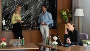 Een hotellobby met een marmeren muur, een receptiebalie en een zithoek met een laptop en een bloevas.