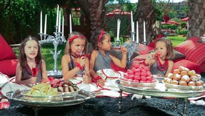 Een groep kinderen zit buiten aan een tafel met eten en drinken, omringd door groen en een decoratieve kandelaar.