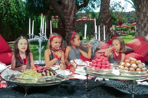 Un grupo de niños sentados al aire libre con una mesa dispuesta con comida y bebidas, rodeados de follaje y un candelabro decorativo.