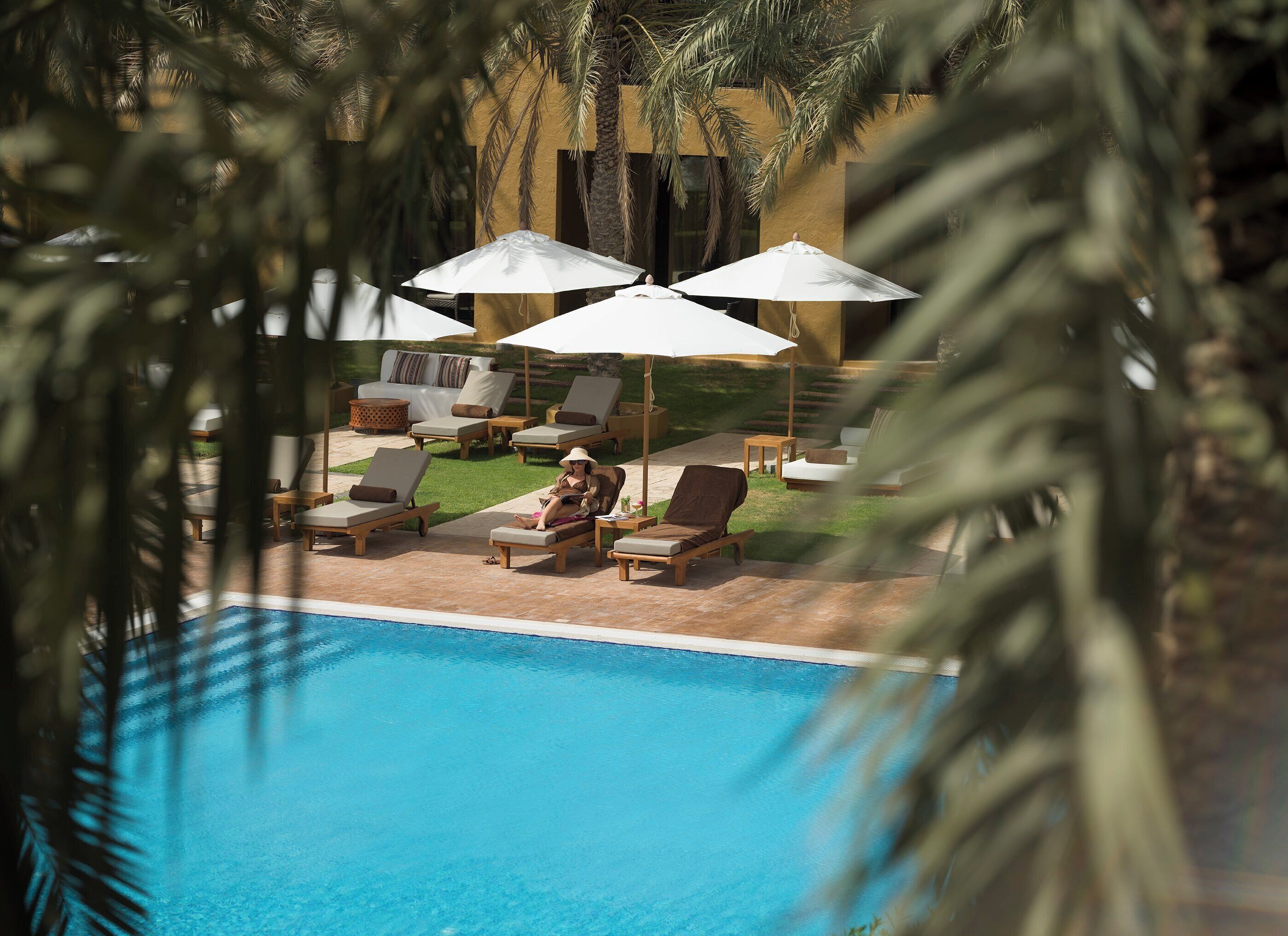 A poolside area with lounge chairs and umbrellas, surrounded by palm trees and a building with a yellow facade.