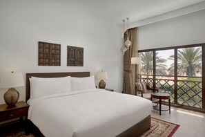 A hotel room with a large bed, two wall-mounted decorative panels, a balcony with a view of palm trees, and a seating area with a table. - Aldhafra Resort, Vignette Collection, an IHG Hotel (Madinat Zayed)
