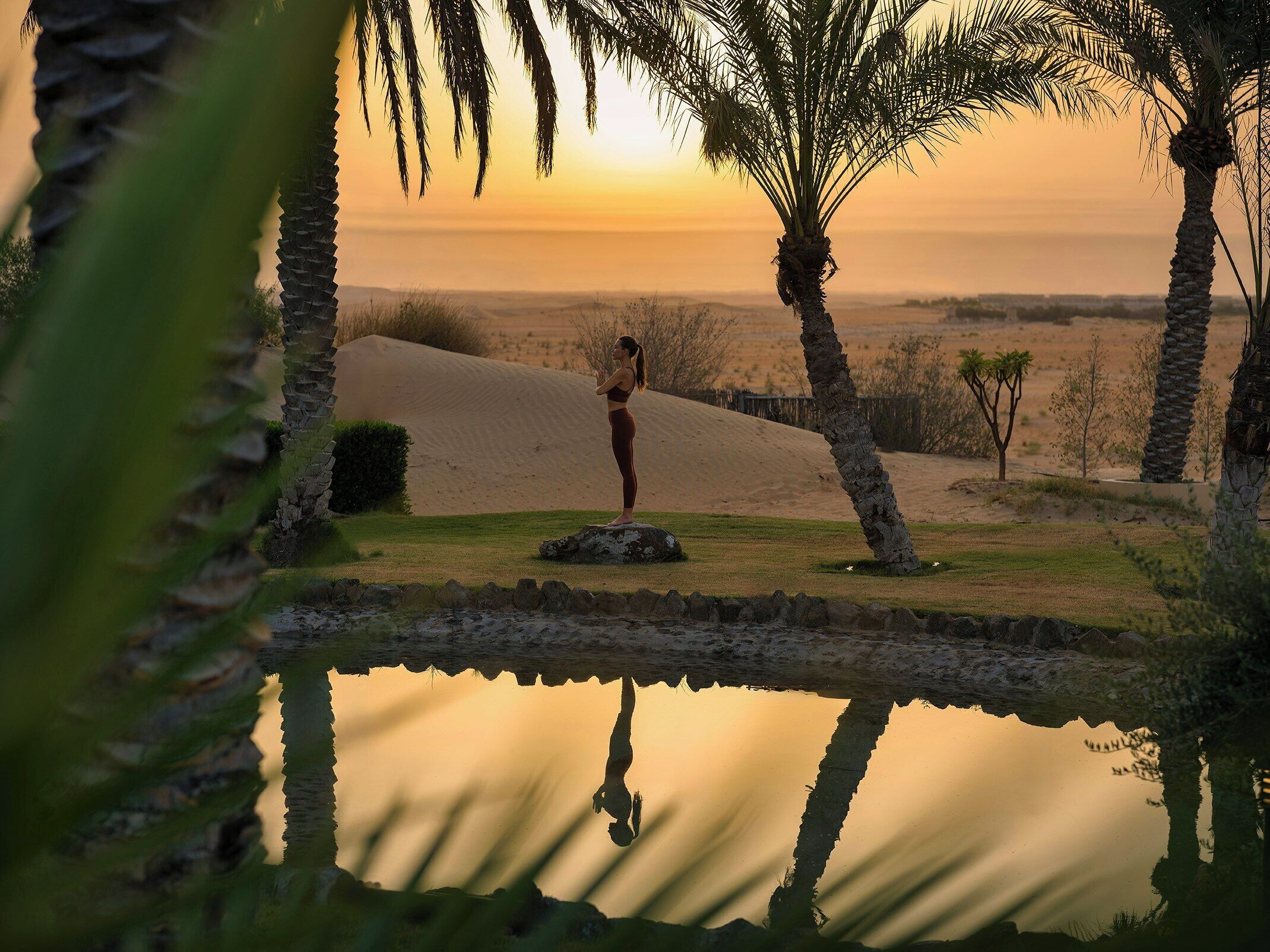 A person meditating by a calm water body with sand dunes and palm trees in the background.