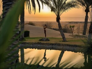 A person meditating by a calm water body with sand dunes and palm trees in the background. - Aldhafra Resort, Vignette Collection, an IHG Hotel (Madinat Zayed)
