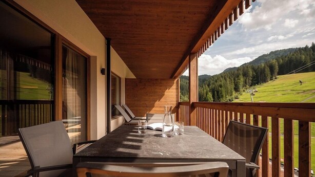 Une terrasse en bois avec une table et des chaises, donnant sur un paysage verdoyant parsemé d’arbres et de collines.