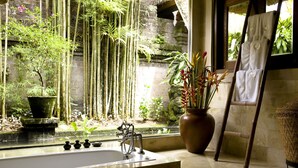 A bathroom with a large bathtub, a stone wall, bamboo plants, and a potted plant with red flowers.