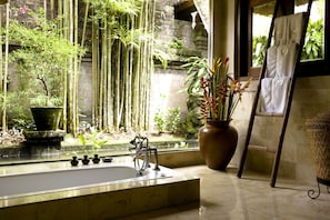 A bathroom with a large bathtub, a stone wall, bamboo plants, and a potted plant with red flowers.