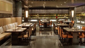Un restaurant au décor moderne, avec une longue table à manger entourée de chaises, un bar avec des étagères remplies de bouteilles, et un chef visible au fond de la salle.