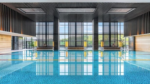 Une piscine intérieure offrant une vue dégagée sur l’extérieur grâce à de grandes fenêtres, un design moderne au niveau du plafond et un sol carrelé.