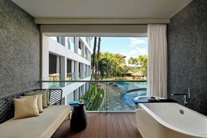 A modern bathroom with a freestanding bathtub, a glass-enclosed window, and a view of a swimming pool and palm trees. - The Stones Hotel Legian Bali Autograph Collection (Legian)