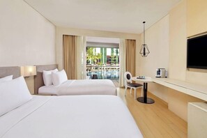 A hotel room with two beds, a TV, a small table, and a view of a pool and palm trees. - The Stones Hotel Legian Bali Autograph Collection (Legian)