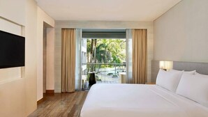 A hotel room with a large bed, a flat-screen TV, a balcony with curtains, and a view of greenery.