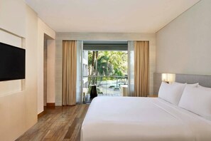 A hotel room with a large bed, a flat-screen TV, a balcony with curtains, and a view of greenery. - The Stones Hotel Legian Bali Autograph Collection (Legian)