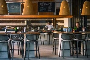 A restaurant with a bar area, bar stools, and a chalkboard menu listing items like salmon and red snapper. - The Stones Hotel Legian Bali Autograph Collection (Legian)