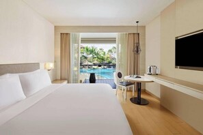 A hotel room with a large bed, a desk with a chair, a TV, and a view of a pool and palm trees. - The Stones Hotel Legian Bali Autograph Collection (Legian)