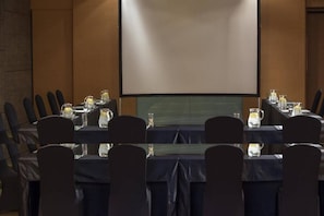 A conference room with a large screen, tables with chairs, and water pitchers. - The Stones Hotel Legian Bali Autograph Collection (Legian)