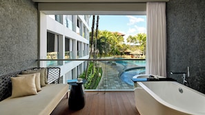 Une salle de bain moderne avec une baignoire, un coin salon avec des coussins et une vue sur une piscine et un bâtiment.