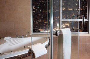 Une salle de bain moderne avec une baignoire, une douche à l’italienne en verre et une vue sur le paysage urbain illuminé la nuit.