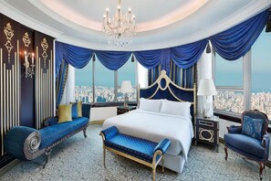A luxurious bedroom with a large bed, a bench, and a chair, featuring a city view through large windows. - Al Habtoor Grand Hotel Beirut (Sin El Fil)