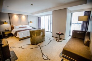 Una habitación de hotel moderna con una cama grande, una zona de estar con sofá y sillón, una mesita redonda y un ventanal con vistas a la ciudad.