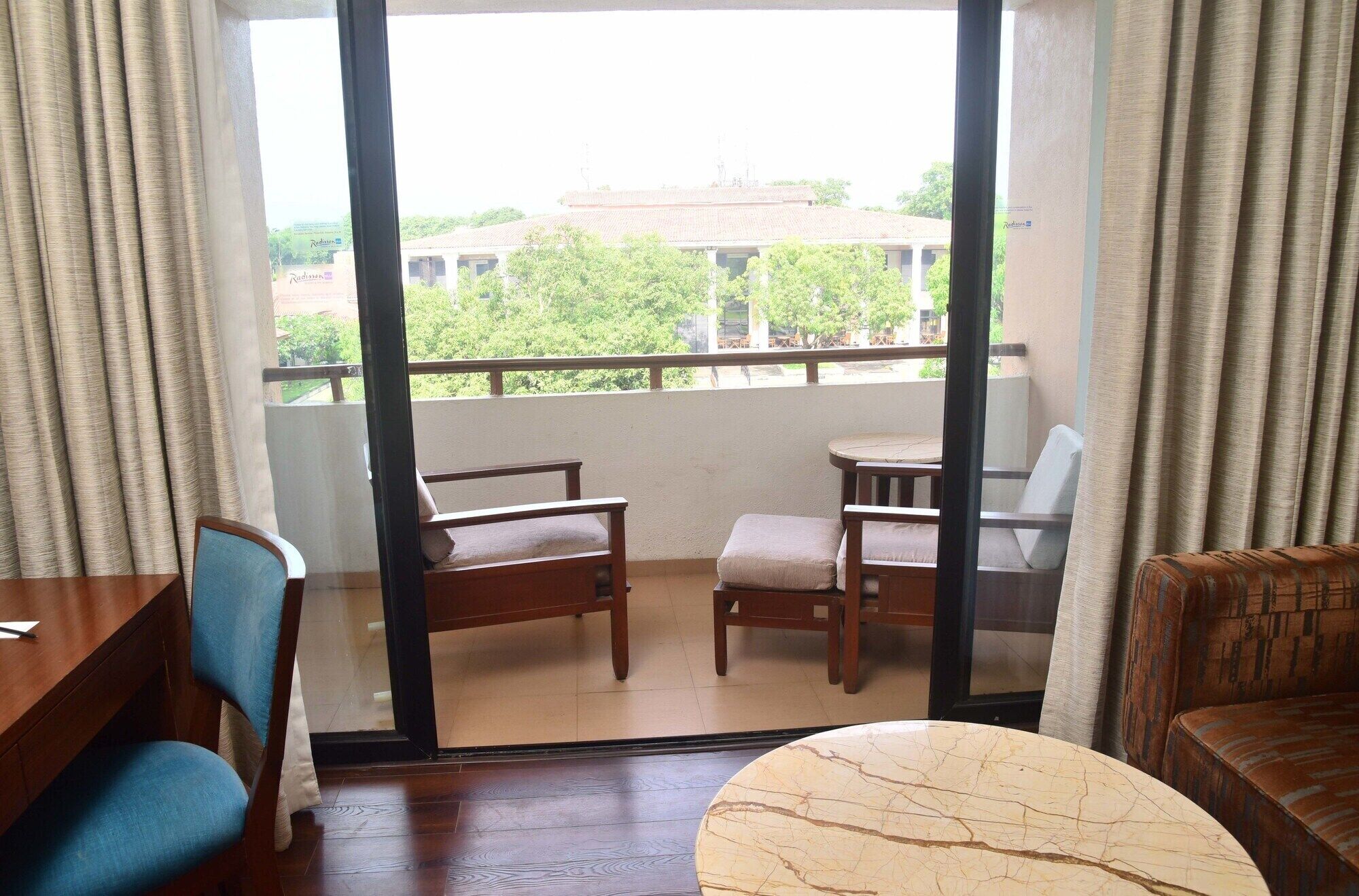 A balcony with a table and chairs, a view of greenery and buildings, and a window with curtains.