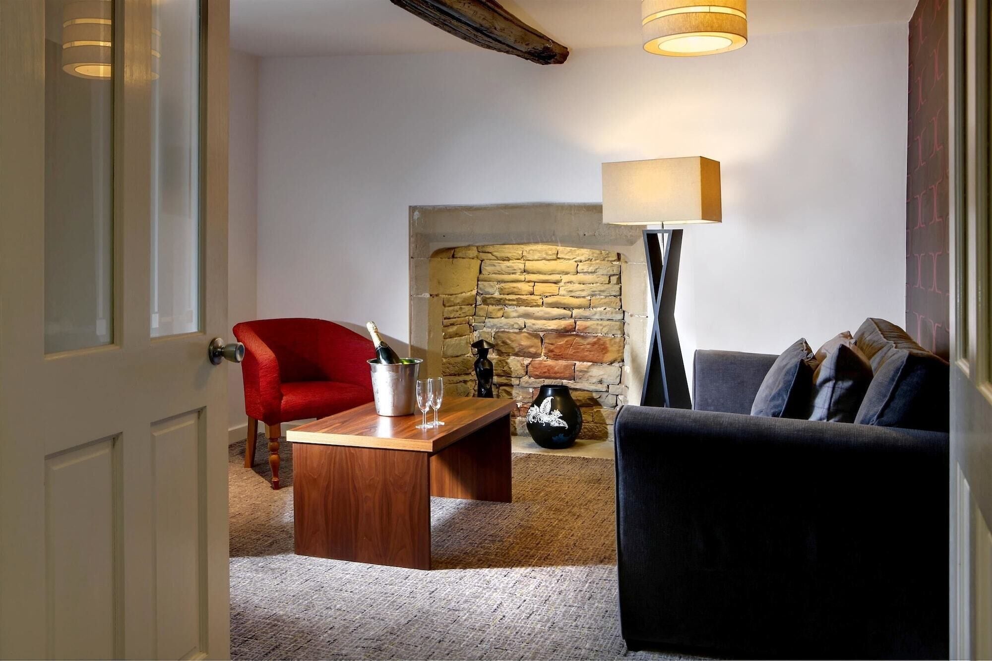 A cozy living room with a stone fireplace, a red armchair, a grey sofa, and a wooden coffee table with a bottle of champagne and glasses.