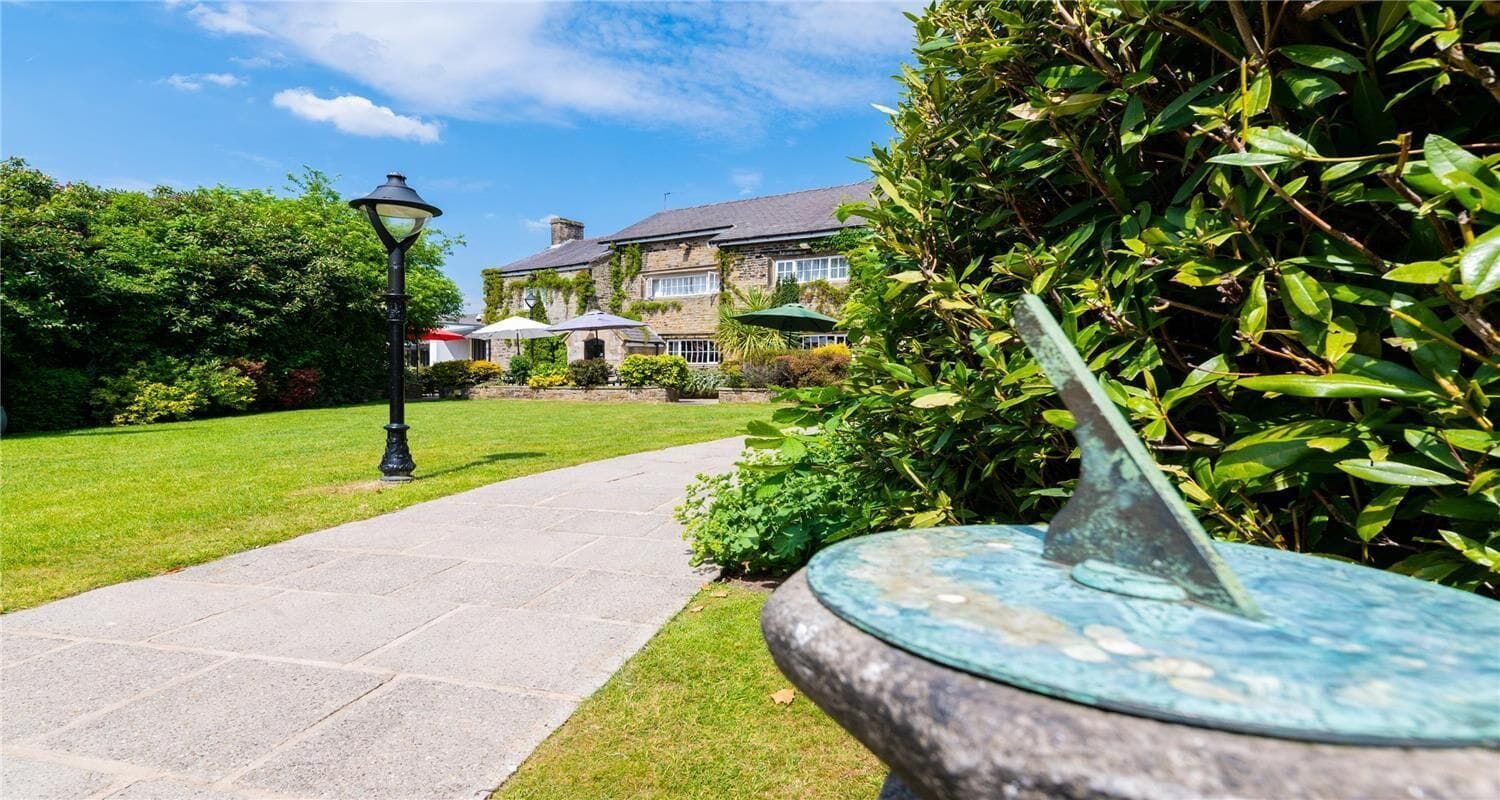 A pathway leading to a stone building with a garden and a sundial.