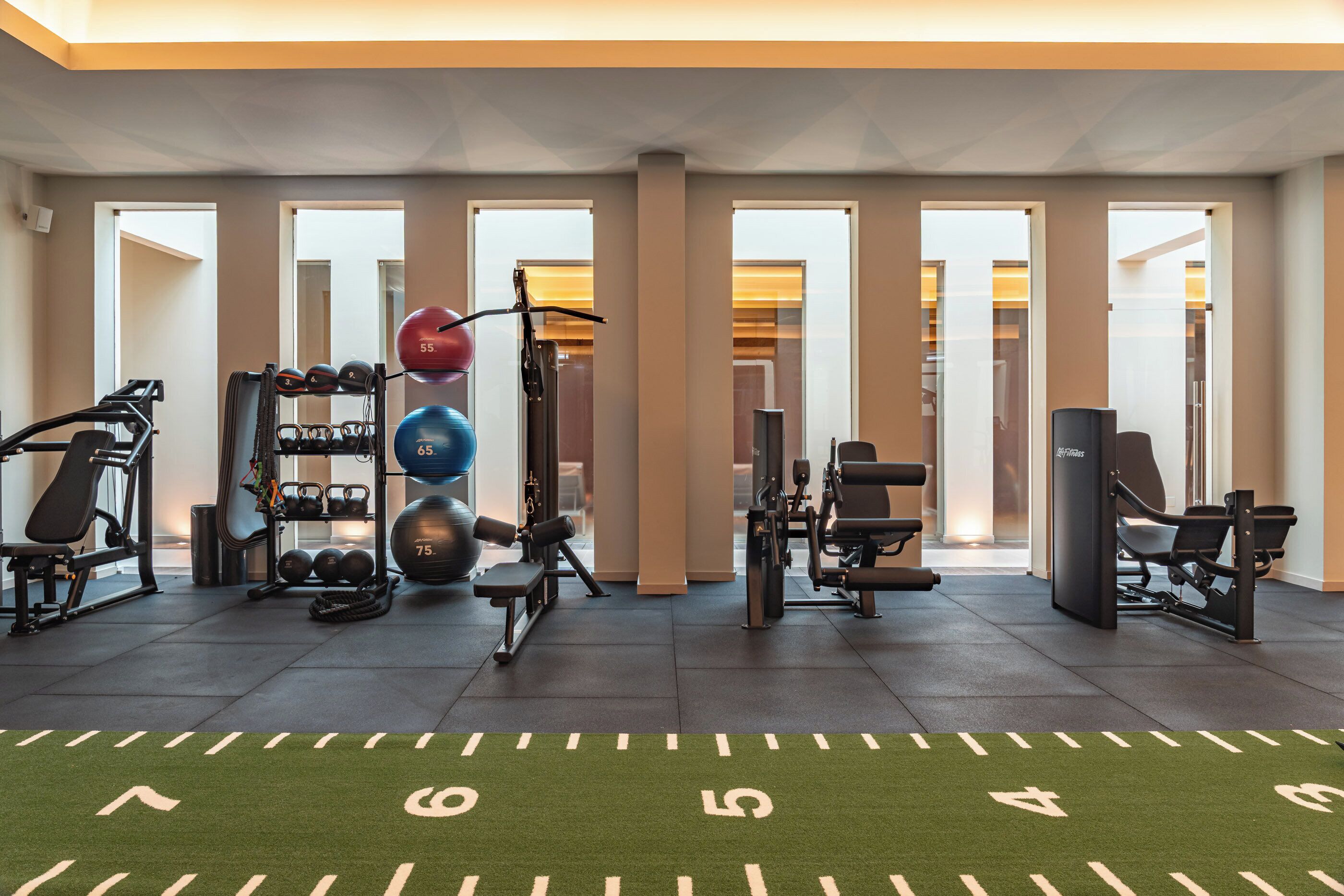 Een fitnessruimte met trainingsapparatuur, een groene grasmat met cijfers en grote ramen.