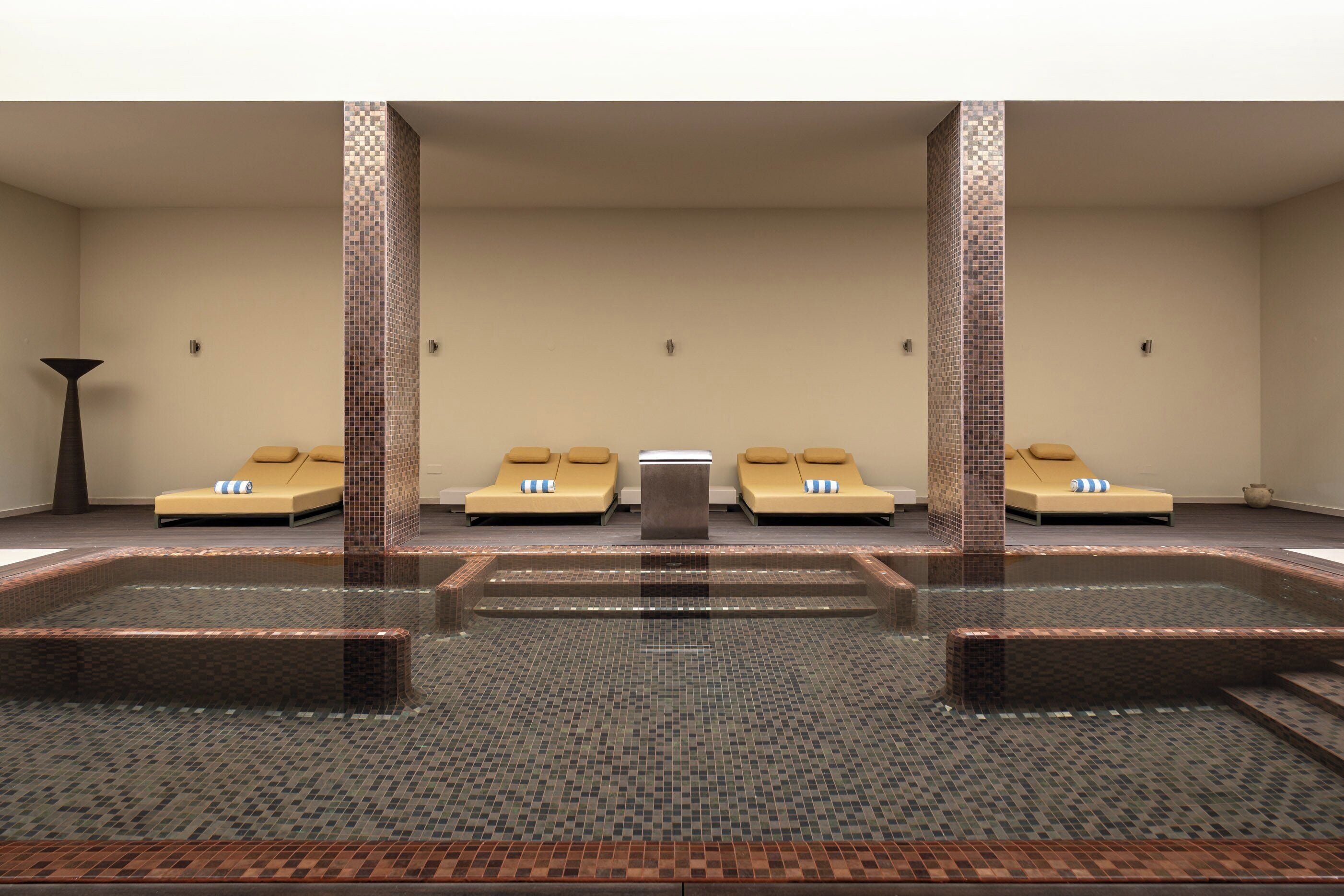 Een wellnessruimte met een centrale whirlpool, vier beige ligbedden en een betegelde vloer.