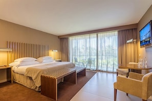 A hotel room with a large bed, a bedside table, a desk with a chair, and a large window with curtains.