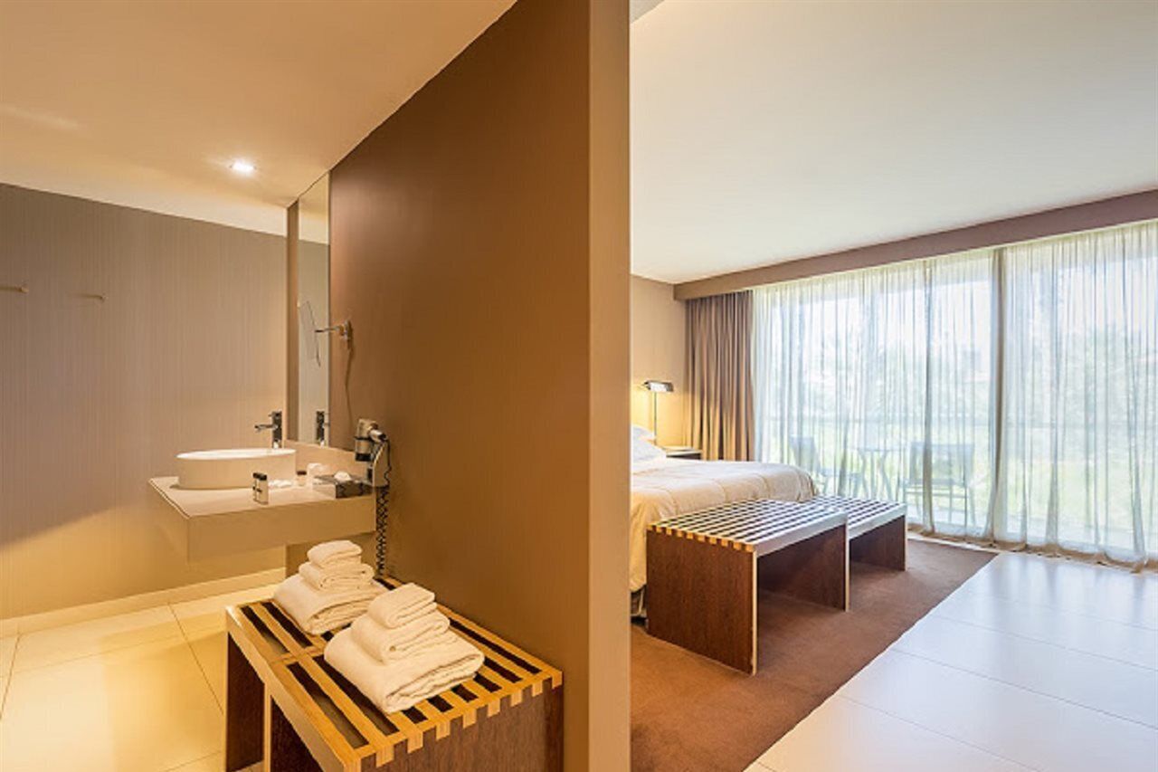 Een moderne hotelkamer met een badkamer, voorzien van een wastafel, spiegel en een houten bankje met handdoeken. De kamer heeft een bed, een nachtkastje en een groot raam met gordijnen.