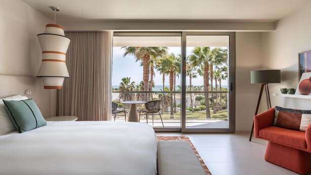 Hotelkamer met een groot bed, zithoek met een tafel en stoelen, rode bank, balkon met uitzicht op palmbomen en de oceaan, en een hanglamp.