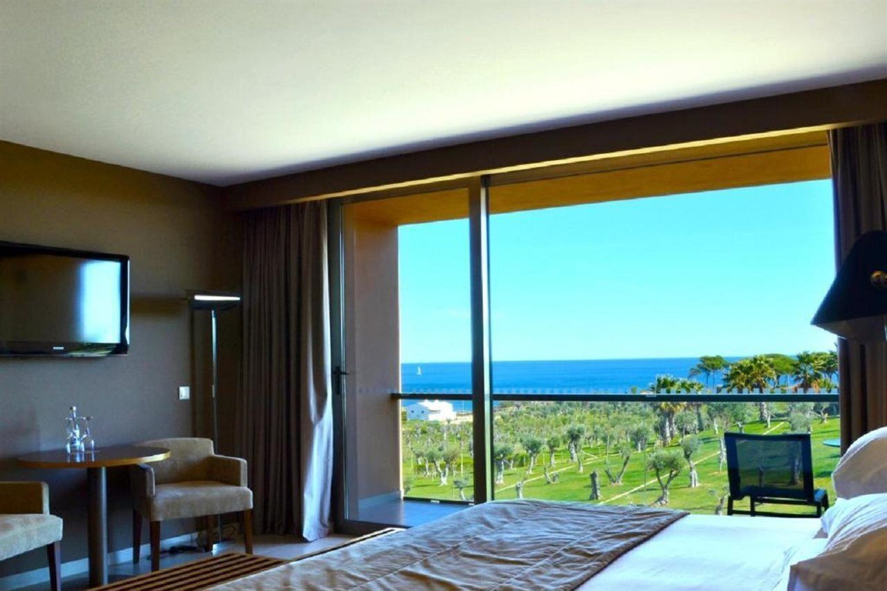 A hotel room with a large window, a bed, a television, and a seating area.
