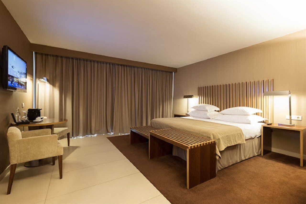 Quarto de hotel com cama, escrivaninha, cadeira, televisão e uma janela grande com cortinas.