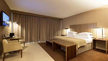 Quarto de hotel com cama, escrivaninha, cadeira, televisão e uma janela grande com cortinas.
