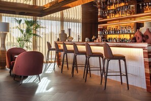 A bar area with wooden bar stools, a potted plant, and a variety of bottles on shelves. - Kimpton Atlântico Algarve, an IHG Hotel (Albufeira)