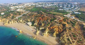 Vista aérea de una zona costera con una playa, acantilados rocosos y una zona residencial al fondo.
