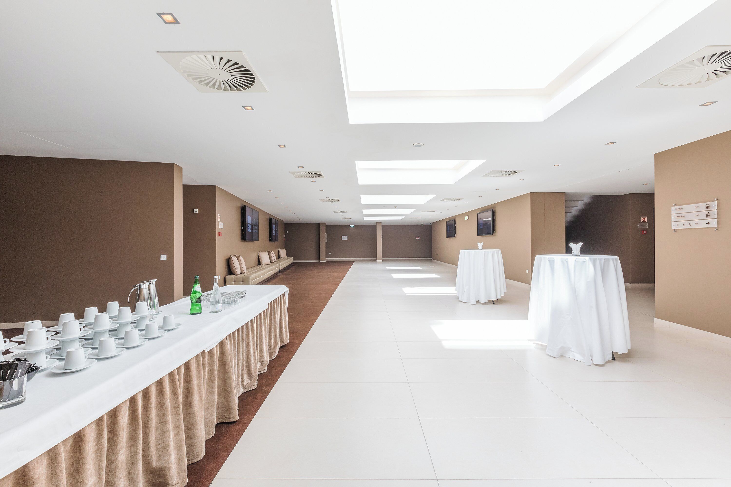 Een ruime hotelgang met een lange receptiebalie, witte tafellakens en een hoog plafond met dakramen.