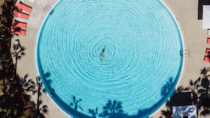 Vue aérienne d’une piscine circulaire où une personne nage, entourée de chaises longues rouges et de palmiers.