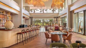 Um amplo saguão de hotel com um bar central, áreas de estar e grandes janelas com vista para o exterior.