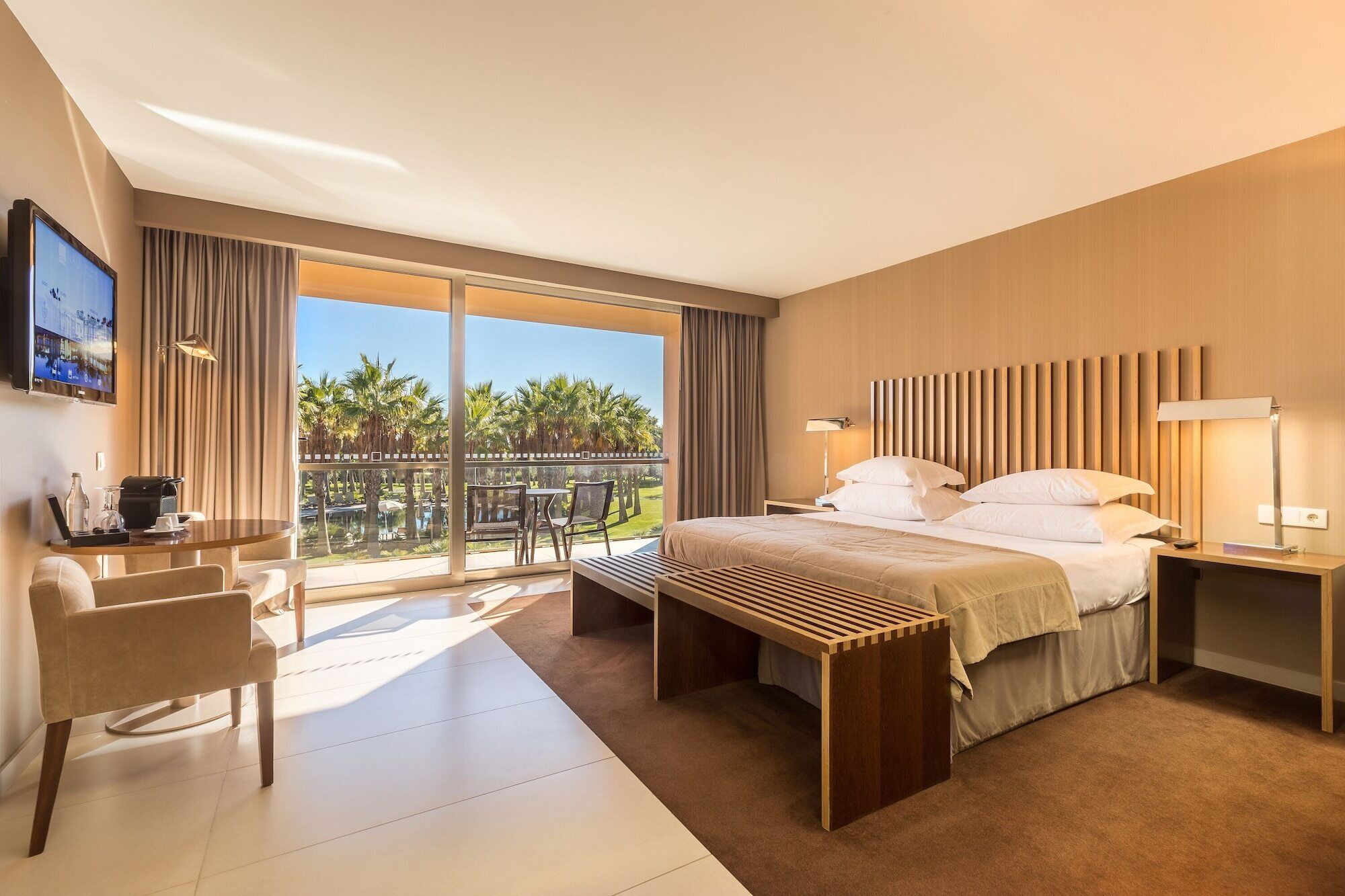 Quarto de hotel com cama, mesas de cabeceira, escrivaninha, cadeira e uma janela grande com vista para palmeiras.