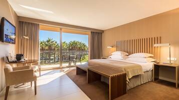 Quarto de hotel com cama, mesas de cabeceira, escrivaninha, cadeira e uma janela grande com vista para palmeiras.
