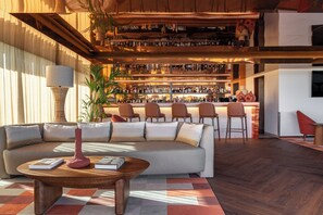 A modern bar area with a sofa and chairs in the foreground, a bar counter with numerous bottles, and a large plant. - Kimpton Atlântico Algarve, an IHG Hotel (Albufeira)