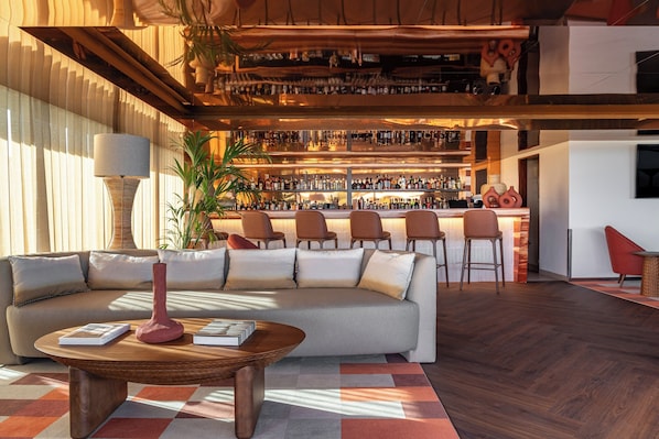 Un espace bar moderne avec un canapé et des fauteuils au premier plan, un comptoir de bar garni de nombreuses bouteilles et une grande plante.