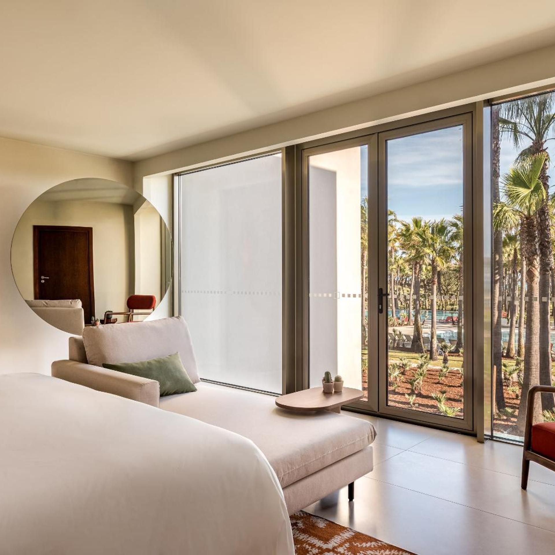 Een moderne hotelkamer met een groot bed, een bank, een kleine tafel en een grote schuifglazen deur die leidt naar een balkon met palmbomen.