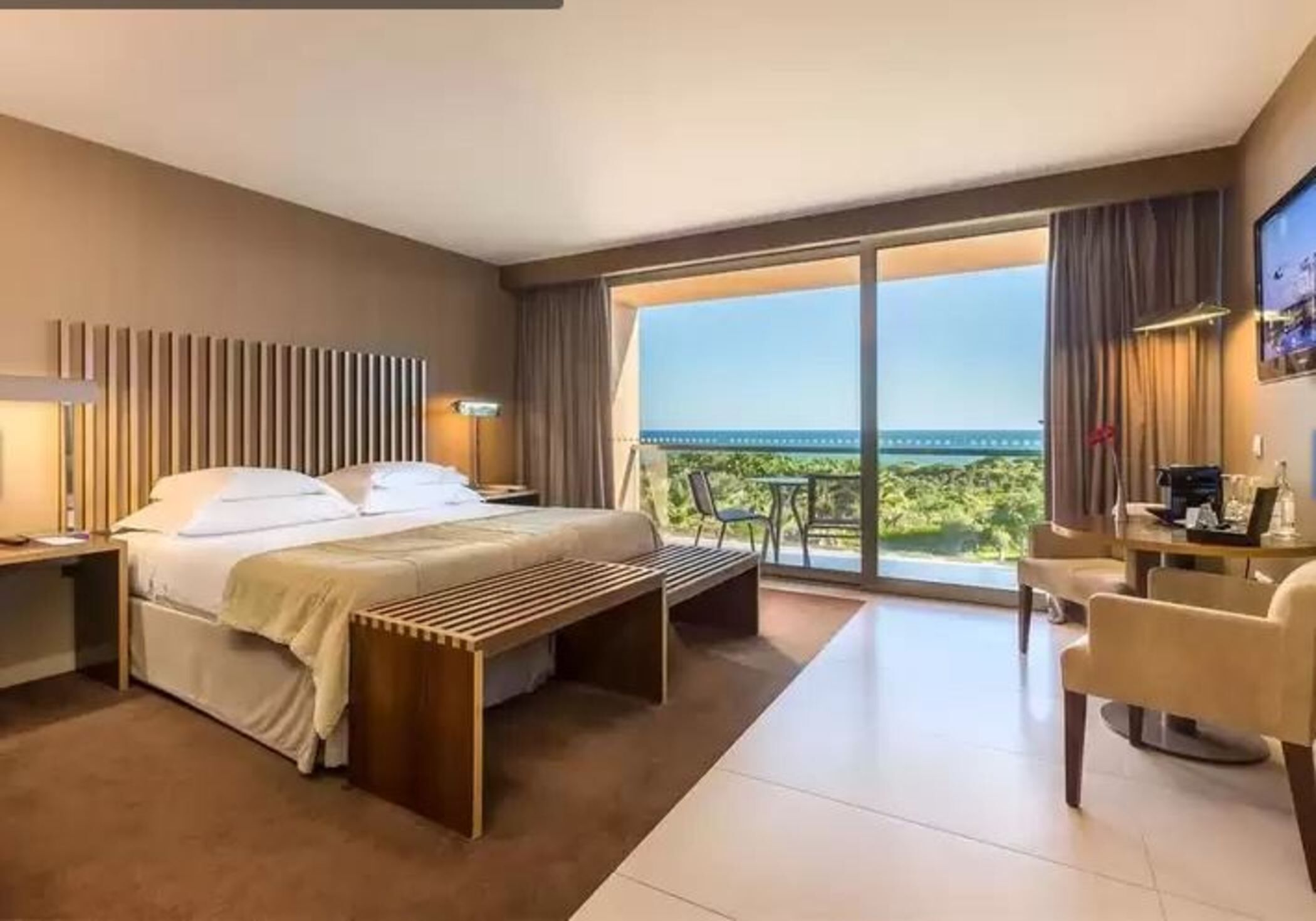 Hotelkamer met een bed, nachtkastje, lamp, bankje en uitzicht op het groen door grote ramen.