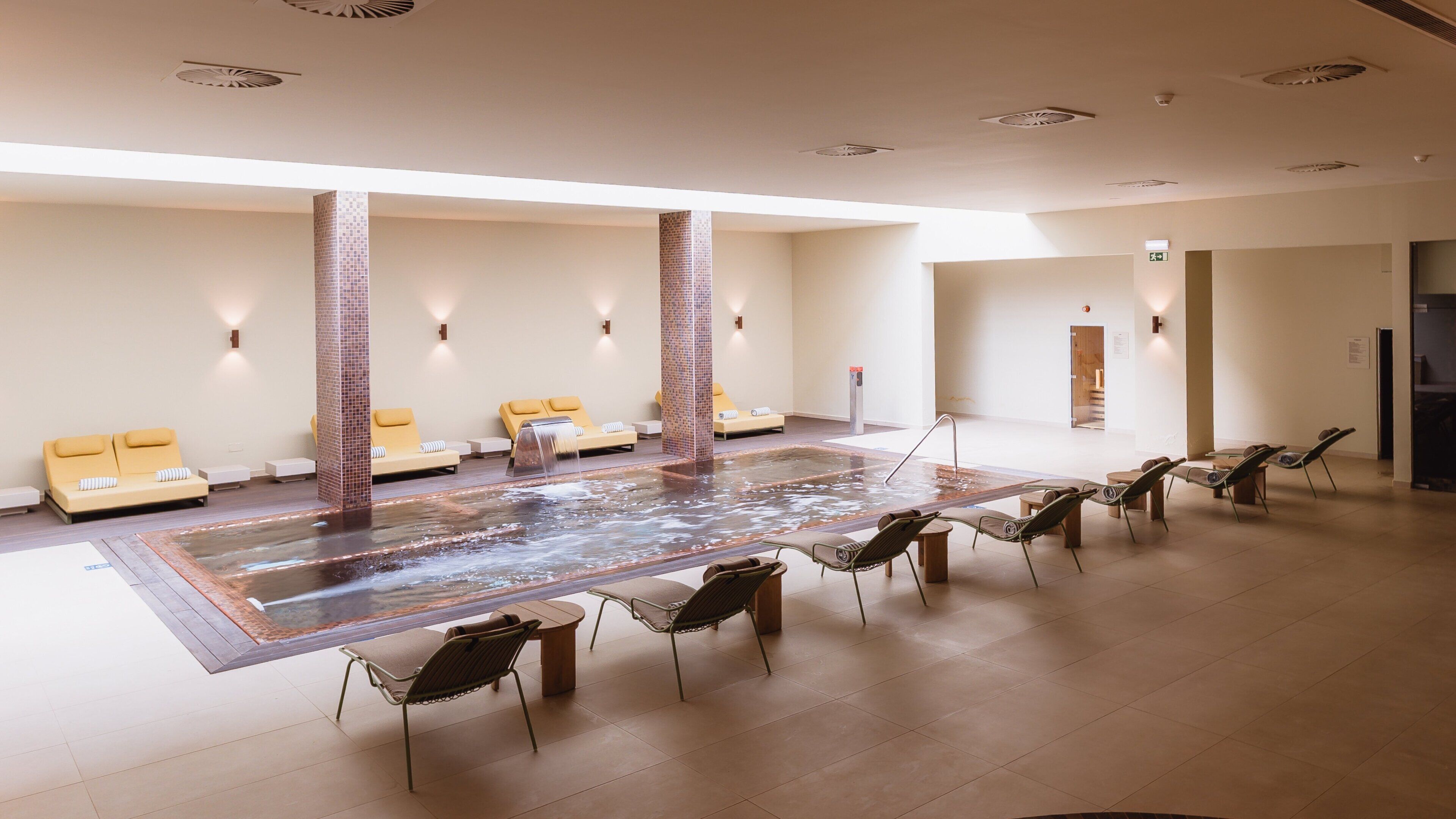Een wellnessruimte met een jacuzzi, lounge fauteuils en een zithoek met gele kussens.