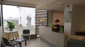 A reception area with a counter, chairs, a table, and a view through a window. - Brit Hotel Hermes (Couchey)