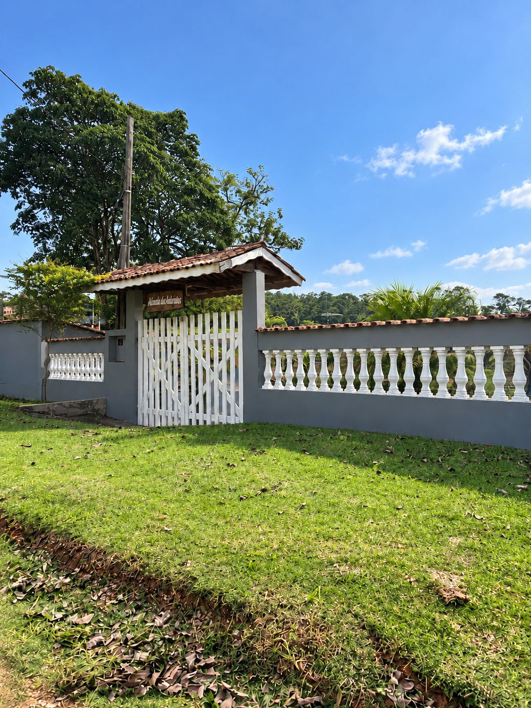 A gated property with a white picket fence, a small building with a tiled roof, and a grassy lawn.
